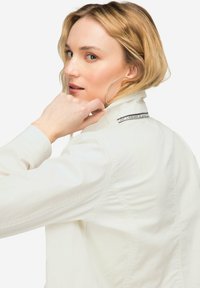 Weiße Denimjacke mit Nähdetails und einem Etikett am Kragen. Das Model hat schulterlanges Haar und trägt große Creolen.