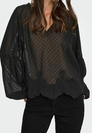 Woman wearing a sheer black long-sleeve blouse with dotted texture and floral embroidery paired with black pants.