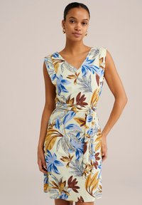 Kleid mit floralem Muster, das blaue, braune und grüne Blätter auf einem hellgelben Hintergrund zeigt. V-Ausschnitt mit Rüsche an den Schultern und einem Bindegürtel.