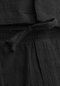 Un tissu noir texturé présente un motif plissé, avec un détail de taille cintrée et des variations subtiles de tissage. Convient pour une tenue décontractée.