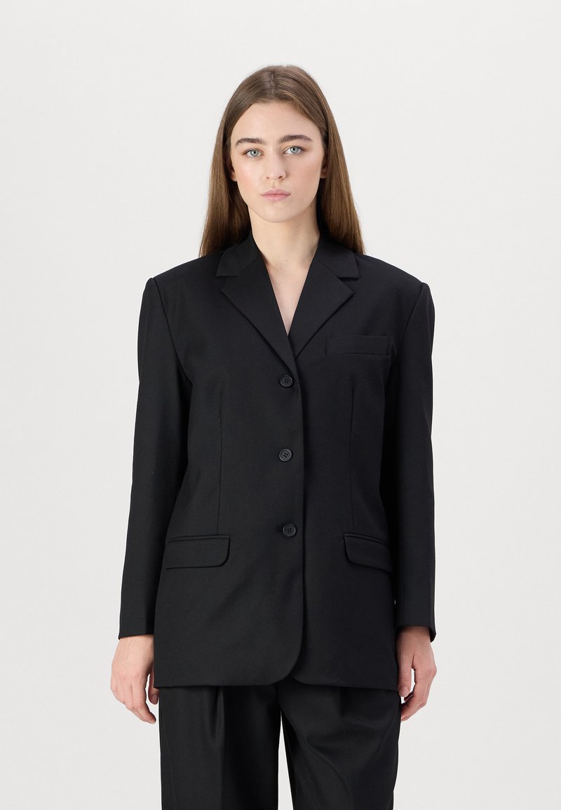 Svart skreddersydd blazer med strukturerte skuldre, hakelapper, fire knapper og to frontlommer. Glatt stoff med en lett glans.