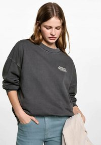 Sivý oversized sveter s rebrovaným golierom a vyšitým textom. Dlhé rukávy, jemná textura. Skombinovaný s svetlomodrými nohavicami.