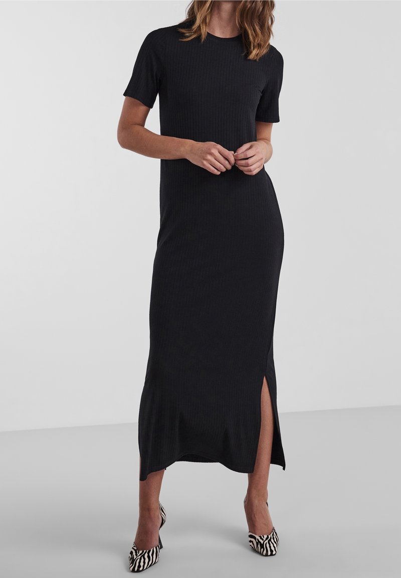 Femme portant une robe midi noire côtelée à manches courtes avec une fente latérale et des talons hauts à imprimé zèbre, debout devant un fond uni.