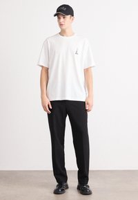 Weißes T-Shirt mit kurzen Ärmeln und kleinem schwarzem Logo, kombiniert mit schwarzen Hosen und schwarzen Schuhen. Die Mütze ist schwarz mit weißer Schrift.