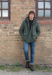 Grüne, gesteppte Kapuzenjacke mit zwei Vordertaschen, schwarzer Rollkragenpullover, blaue Jeans und braune Schnürstiefel vor einer Backsteinmauer.