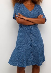 Kvinna med lockigt hår klädd i en blå, kortärmad klänning med knappar framtill och ett litet blommönster, stående med armarna i kors.