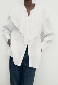 Chemise blanche avec un devant à volants et un col mandarin, dotée de manches longues et d'une coupe décontractée. Tissu texturé et fermeture par boutons.