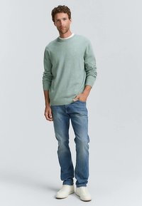 Minzgrüner Pullover mit Rundhalsausschnitt, getragen über einem weißen T-Shirt, kombiniert mit hellblauen Jeans und weißen Turnschuhen.