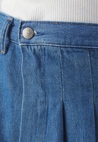 Blå denimkjol med veck, silverfärgad knapp och kontrasterande orange sömmar, vilket visar en texturerad tyg.