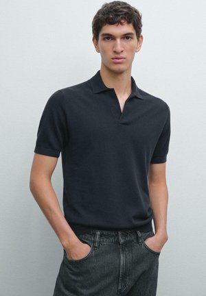Dunkles Poloshirt aus weichem Stoff mit kurzem Kragen und kurzen Ärmeln, kombiniert mit grauen Denim-Hosen. Das Model steht mit den Händen in den Taschen.