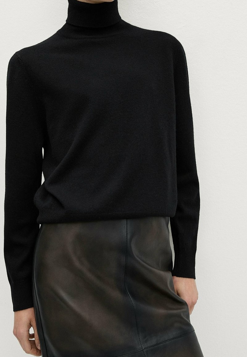 Personne portant un pull à col roulé noir rentré dans une jupe en cuir noire taille haute, sur un fond clair uni.