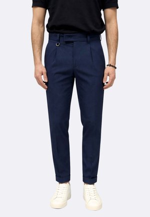 Uomo che indossa pantaloni blu navy su misura con orli risvoltati, una camicia nera infiocchettata, sneakers bianche e un braccialetto nero al polso destro.