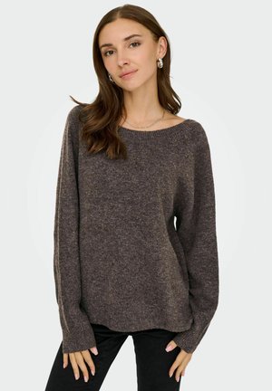 Femme aux longs cheveux bruns portant un pull en maille ample gris foncé et un pantalon noir sur un fond clair uni.