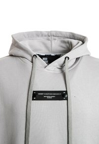 Sudadera gris claro con cordón en el cuello, tejido texturizado y una etiqueta de marca negra en el pecho con texto blanco.