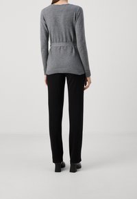 Kobieta stojąca tyłem, ubrana w szary sweter z długim rękawem i paskiem, czarne proste spodnie oraz czarne buty na obcasie, na jednolitym tle.