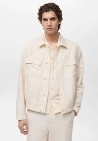 Hellbeige Jacke mit langen Ärmeln, vier Taschen vorne und Knopfverschlüssen; aus einem strukturierten Stoff gefertigt, über einem passenden Outfit getragen.