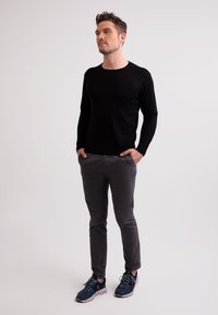 Schwarzer langärmeliger Pullover, graue slim-fit Hose und marineblaue Sportschuhe. Das Model steht mit den Händen in den Taschen vor einem hellen Hintergrund.