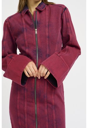 Femme portant une veste rouge à fermeture éclair à manches longues avec de larges poignets, les mains jointes près de la taille devant un fond uni.