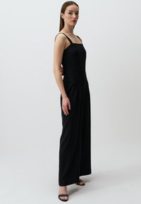 Zwarte jumpsuit met dunne bandjes, een vierkante halslijn en wijde benen. Glad materiaal met een aansluitende lijfje en plooien bij de taille. Minimalistisch ontwerp.