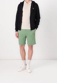 Muž v černé zipové mikině, béžovém polo tričku, zelených šortkách se žlutým logem, bílých ponožkách a bílých teniskách na jednobarevném pozadí.