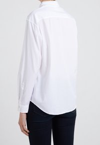 Camicia bianca a maniche lunghe con bottoni, dotata di yoke posteriore, orlo arrotondato e polsini; tessuto liscio con una vestibilità rilassata. Indossata con jeans scuri.