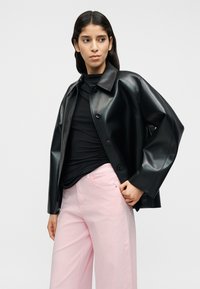 Μαύρο τζάκετ από συνθετικό δέρμα με γιακά, κουμπιά μπροστά και φαρδιά μανίκια, συνδυασμένο με ανοιχτό ροζ παντελόνι φαρδιάς γραμμής.