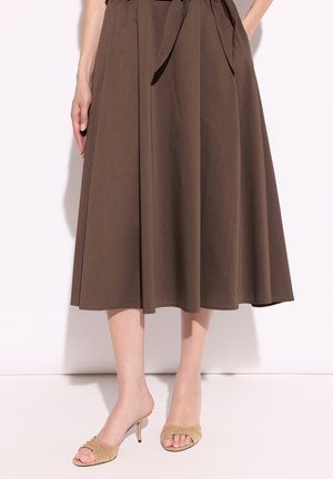 Kobieta w brązowej spódnicy midi z wiązanym paskiem w talii i beżowych sandałach na obcasie z odkrytymi palcami, stojąca na prostym jasnym tle.