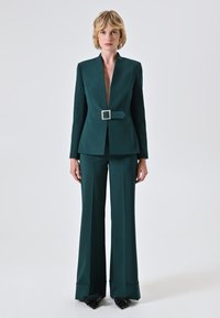 Completo sartoriale verde scuro con blazer a profonda scollatura a V, cintura in vita e pantaloni a gamba larga con una leggera svasatura all'orlo. Scarpe nere.