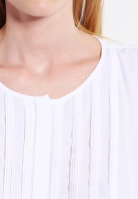 White blouse with a scooped neckline, featuring vertical pleats, light, airy texture, and a button closure. Subtle and clean design elements.