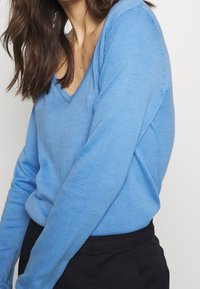 Blauer, leichter Pullover mit V-Ausschnitt. Verfügt über lange Ärmel und gerippte Akzente am Ausschnitt und an den Bündchen. Glatte Textur, taillierte Passform.