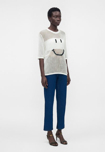 Γυναίκα που φοράει λευκή διάφανη μπλούζα με μαύρο χαμογελαστό πρόσωπο, μπλε παντελόνι με ίσια μπατζάκια και μαύρα ψηλοτάκουνα σανδάλια με λουράκια στον αστράγαλο.