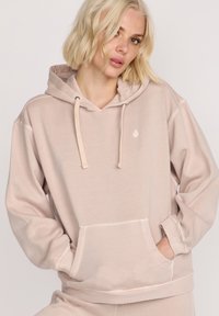 Lys beige hoodie med snøre, frontlomme og ribbede manchetter. Har et lille logo på brystet. Blødt, afslappet stof og løs pasform.