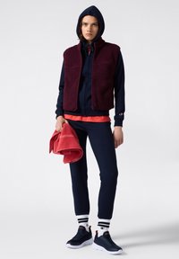 Conjunto deportivo que incluye un chaleco de forro polar color burdeos oscuro, sudadera con capucha azul marino y pantalones a juego. Zapatillas negras con suelas blancas y detalles rojos. Toalla roja en la mano.