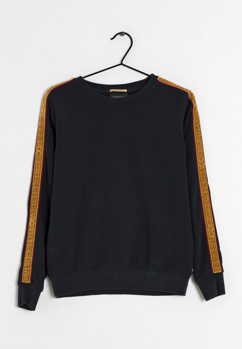 Sweat-shirt noir à manches longues avec col rond, suspendu sur un cintre noir, avec des rayures jaunes à motifs le long des manches.