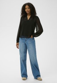 Zwarte, transparante blouse met lange mouwen en een v-hals, gecombineerd met lichtblauwe wijde jeans en beige schoeisel.