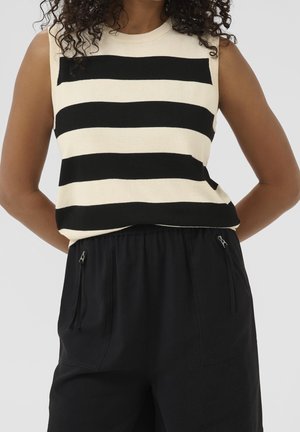 Top sin mangas beige con amplias franjas horizontales negras combinado con pantalones negros holgados con bolsillos con cremallera.