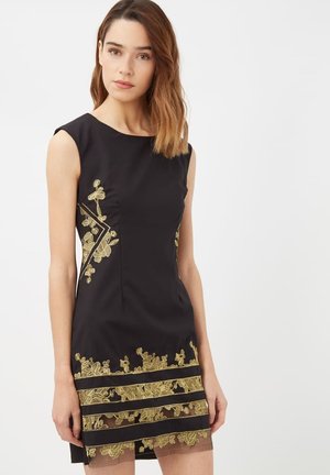 Mujer vestida con un vestido negro sin mangas, con bordados florales dorados y rayas horizontales transparentes cerca del dobladillo, de pie frente a un fondo claro y liso.