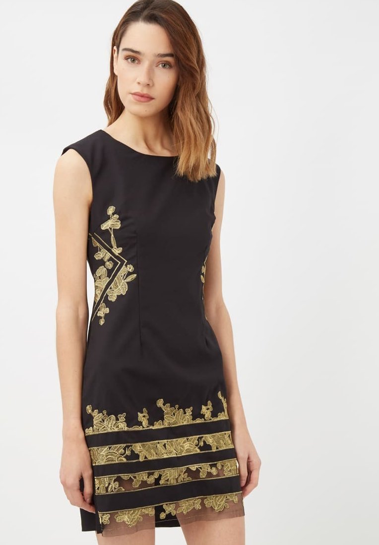 Mujer vestida con un vestido negro sin mangas, con bordados florales dorados y rayas horizontales transparentes cerca del dobladillo, de pie frente a un fondo claro y liso.