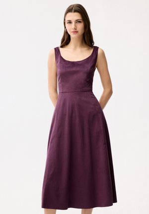 Młoda kobieta z długimi brązowymi włosami, ubrana w bezrękawnikową, dopasowaną fioletową sukienkę midi z dekoltem w szpic, stojąca na białym tle.