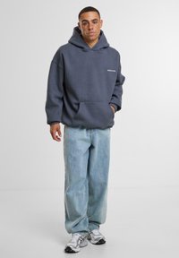 Gray oversized hoodie with a front pocket and white logo detail, paired with light blue wide-leg jeans and silver sports shoes.