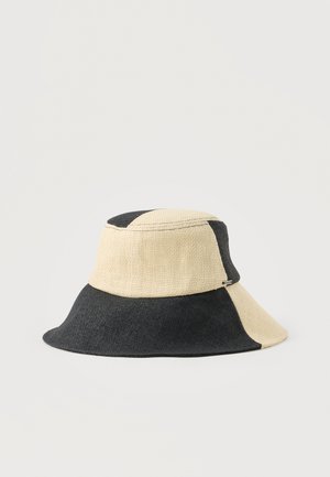 Bézs és fekete színű, széles karimájú, foltdíszes mintával és szőtt textúrával rendelkező vödörkalap, egyszerű világos háttéren bemutatva.