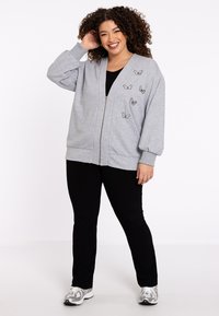 Grau gestrickte Sweatjacke mit Ballonärmeln, Schmetterlings-Stickerei auf der Vorderseite, kombiniert mit schwarzen Hosen und Sneakers. Glatte Stofftextur.