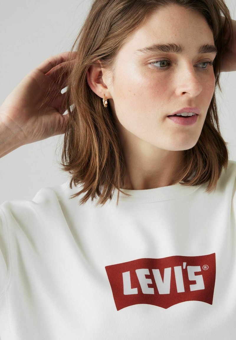 Vit sweatshirt med en stor röd Levi's-logotyp. Modellen har axellångt brunt hår och bär små guldörhängen.