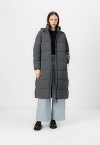 Lang grå quiltet frakk med hætte, der har en frontlynlås, en kviltet design og sidelommer. Sammen med lyseblå vide jeans.