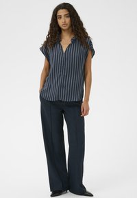 Tengerészkék csíkos rövid ujjú ing gallérral, tengerészkék bő szárú nadrággal és hegyes orrú fekete cipővel viselve. Sima anyag és laza szabás.