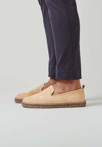 Chaussures slip-on en suède beige avec un bout arrondi et une semelle en corde de jute, dotées d'un petit talon et d'un logo embossé sur le côté.