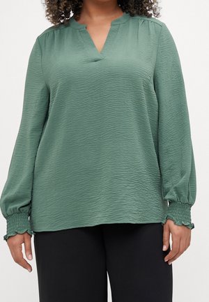 Nainen, jolla on päällään teksturoitu salviavihreä pitkähihaisen paita, jossa on smokatut hihansuut, ja mustat housut. Kuvassa näkyy keskirinnasta lantiolle asti.