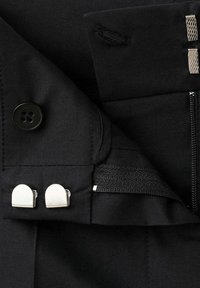 Tissu noir avec attaches à crochets et barres argentées, fermeture éclair et fermeture par bouton sur le vêtement, montrant un détail de la ceinture intérieure.