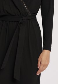 Robe noire à manches longues avec une ceinture à nouer à la taille et de petites clous métalliques le long de l'encolure, une main reposant sur le côté.