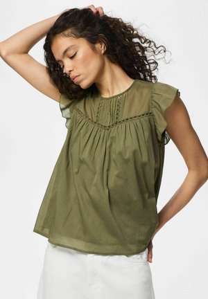 Vrouw met krullend haar die een olijfgroene mouwloze blouse met ruches en een witte broek draagt, poseert met één arm omhoog en ogen dicht.